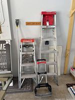 Photo showing three aluminum ladders and one folding hand cart, all standing against a wall indoors.