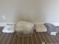 Four folded bedding items arranged on carpet: stack with white king sheets, beige Vera Wang queen sheets and pillowcases, and gray sheets in plastic wrap.