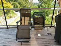 Pair of folding outdoor reclining chairs on wooden deck with glass balcony rail overlooking greenery.