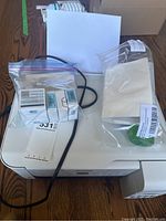 Epson ET-2720 white WiFi printer with control panel and visible ink tanks from front and side angles, accompanied by power cable and sealed ink bottles in bags.