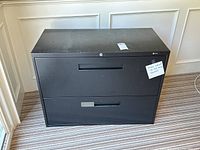 Front view of a black metal filing cabinet with two drawers, showing integrated handles and lock on upper drawer, light wear visible on surface.