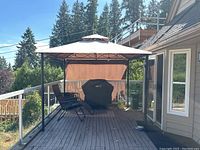 Full view of gazebo on wooden deck with two reclining chairs and covered Weber Q grill, beige canopy and mesh side panel visible.