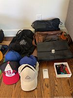Photo of multiple baseball caps in front including a red/white/blue USA cap, beige Adidas cap, two blue caps; several bags behind including brown leather bag, black fabric bag with text, black leather bag