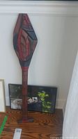 Wood carved paddle standing against the wall with framed print behind it, showing the overall size and display context.