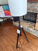 Full view of floor lamp showing wooden tripod base and white cylindrical shade in a room with hardwood floor and stone fireplace.