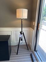 Full view of tall wood tripod floor lamp with white fabric lampshade standing next to window and black cabinet