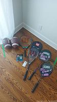 Photo showing two Wilson tennis racquets, two Sugi badminton racquets with covers, two Wilson Grey Cup footballs, and extra grip packs on floor.