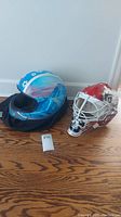 Photo of both helmets side by side on a wooden floor showing the bobsled helmet with its protective bag and the goalie mask