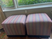 Pair of square ottomans side by side under window, showing striped multi-color fabric upholstery.