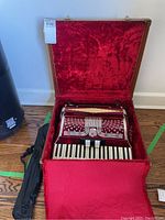 Accordion inside open brown hard case lined with red velvet, black bag for music stand beside it