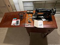 Full view of vintage Singer electric sewing machine mounted on wooden table, showing machine, manual, cords, foot pedal, and accessories spread on table.