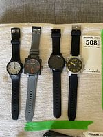 Four watches laid out on white towel including Kenneth Cole Reaction chronograph, Kenneth Cole Reaction black and orange dial, Cookoo smart watch, and Kenneth Cole Reaction watch with yellow numbers. Also visible MaxSold 508 measurement card.