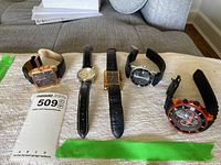 Photo of five men's wristwatches laid out on fabric showing varied styles and cases including rectangular, round, black and gold tones.