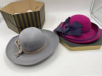 Two vintage felt hats displayed with accompanying striped hatbox. One gray hat with wide brim and beige/light brown striped ribbon bow. One magenta hat with dark purple ribbon bow and turned-up brim.