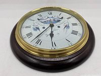 Front view of a round vintage wall clock with a white face, brass bezel, and dark wood mounting, marked G. Stones Blackburn, Roman numerals, decorative birds and a building illustration.