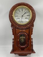 Front view of the wooden regulator wall clock showing round clock face with Roman numerals and brass trim, ornate carved floral woodwork around the frame.