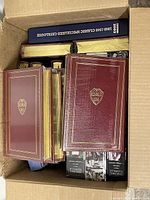 Top view inside box showing multiple burgundy hardcover books with Harvard seals and gold gilded page edges, along with other stacked books and catalogs.