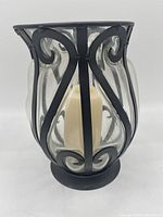 Front view of metal and glass candle holder showing curved glass panels with black metal scrollwork and candle inside.