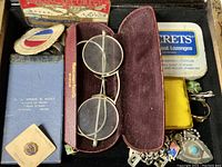 Overview of cigar box contents showing antique spectacles in burgundy case, Sucrets tin, ribboned medals, and other small items.