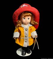 Front view of the doll showing detailed face, red firefighter hat, yellow coat, and black boots on a white stand.