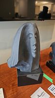 Front view of the black matte bust sculpture showing detailed face and hair, placed on a wooden surface with a TV in the background.