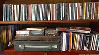 Shelf with multiple CDs in jewel cases and cassettes in cases and a carrying case.
