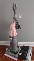Front view of pink Dyson DC07 upright vacuum with power cord wrapped at handle and dusty canister, accompanied by boxed Asthma and Allergy Care Kit on floor