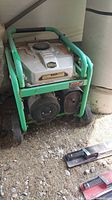 Front and side angled view of the green frame portable generator with visible engine and wheels, set indoors on dirt floor.