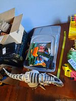 Photo shows large white and black mechanical dinosaur toy on a wooden table with several unopened Hot Wheels cars in original packaging inside a plastic bin in the background.