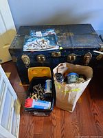 Photo showing a large black storage chest with metal clasps, a black plastic bin filled with assorted Lego Bionicle pieces and containers, and a brown paper bag containing several Bionicle canister containers with graphics.
