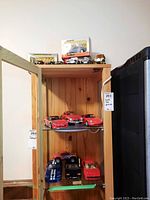 Wooden three-shelf display cabinet holding multiple model cars including unboxed red and blue sports cars and boxed models on top shelf