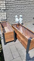 Set of two rectangular wooden planters with black metal corner supports placed outdoors on stone pavement next to garden greenery. Both lined inside with clear plastic liners.