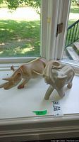 Two statuettes placed side by side on a windowsill with grass visible outside. Elephant has trunk raised and rhinoceros has two horns.