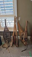 Photo showing two pairs of vintage wooden snowshoes and four vintage wooden lacrosse sticks arranged upright against a wall in a room with carpet and window blinds.