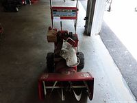 Front view of red and beige Craftsman snow blower showing auger and chute for snow discharge. Machine is equipped with treaded tires.