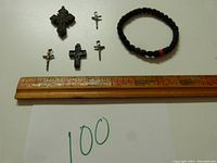 Top view showing all five crosses, three metal and two black plastic, and the textile bracelet laid out next to a ruler for scale.