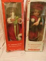 Two boxed 24-inch animated Holiday Caroler figures, one in red dress and one in black coat, both holding candlelight props, with partial box text showing