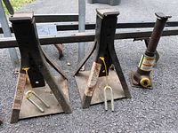 Photo showing two metal jack stands with flat bases and a small hydraulic car lift jack on pavement.