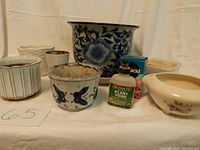 Full view of all planters grouped together on fabric backdrop, including the large blue and white floral ceramic planter, three-footed planter, various smaller planters, and garden products.