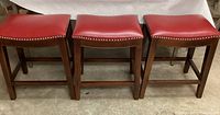 Front view of three red saddle stools with wooden legs and nailhead trim along the seat edges.