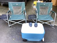 Two green fold-up camping chairs with attached side tables and a blue and white Igloo cooler on wheels with two black camping pillow bags on top, taken outside on pavement.