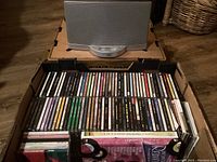 Photo of a cardboard box filled with two rows of assorted music CDs in standard jewel cases, showing a large variety of album spines and titles.