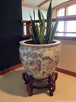 Photo showing a large hand painted ceramic planter pot with snake plant in an interior setting near a window. The planter is on a carved wooden stand.