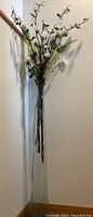 Full view of tall clear glass vase with white artificial flowers set against a corner wall