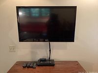 Front view of LG 37-inch flat screen TV mounted on wall mount bracket above wooden surface with Sony Blu-ray DVD player and four remote controls placed on the table.