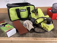 Ryobi electric belt sander with power cord, dust collection bag, green/black storage pouch, new boxed sanding belts and loose sanding belt on workbench.