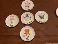 Overview of five Scully and Scully decorative plates with whimsical floral designs on a brown fabric background.