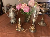 Full view of all items including two candlestick lamps (one with etched glass shade), silver plated hot water pot, tall oil lamp, glass vase with faux pink peonies