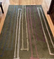 Full view of rectangular wool area rug on hardwood floor, showing dark olive green color with contemporary linear geometric patterns in blues, purples, and white.