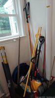 Photo showing various yard tools including rakes, shovels, broom, and the collapsible trash can in corner of room near window.